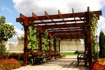 Pergola En Bois Naturel Avec Bancs. L'ensemble Est Dans Un Grand Jardin Fleuri.