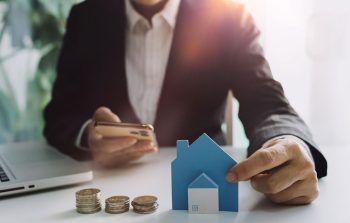 Homme Qui Tient Un Smartphone Et Une Petite Maison Dans Ses Mains. A Coté Des Pièces De Monnaie Et Un Ordinateur